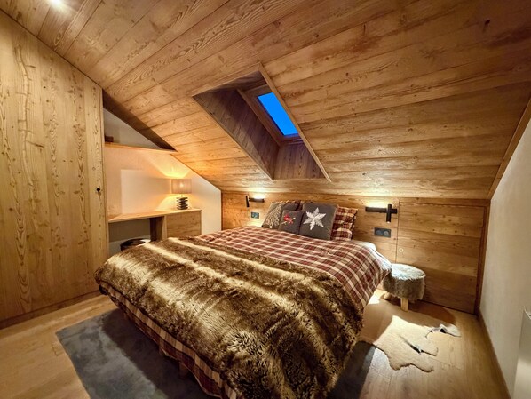 Ein gemütliches Schlafzimmer im Dachgeschoss mit schrägen Wänden, einem Oberlicht, einem Bett mit Felldecke und einem Nachttisch mit Lampe.