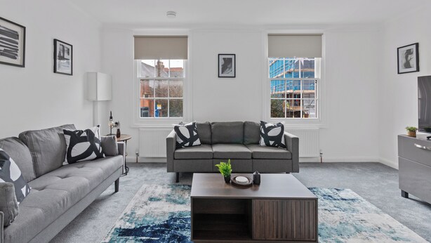 Ein modernes Wohnzimmer mit einem grauen Sofa, einem Couchtisch und einem großen Fenster mit Blick auf ein Gebäude.