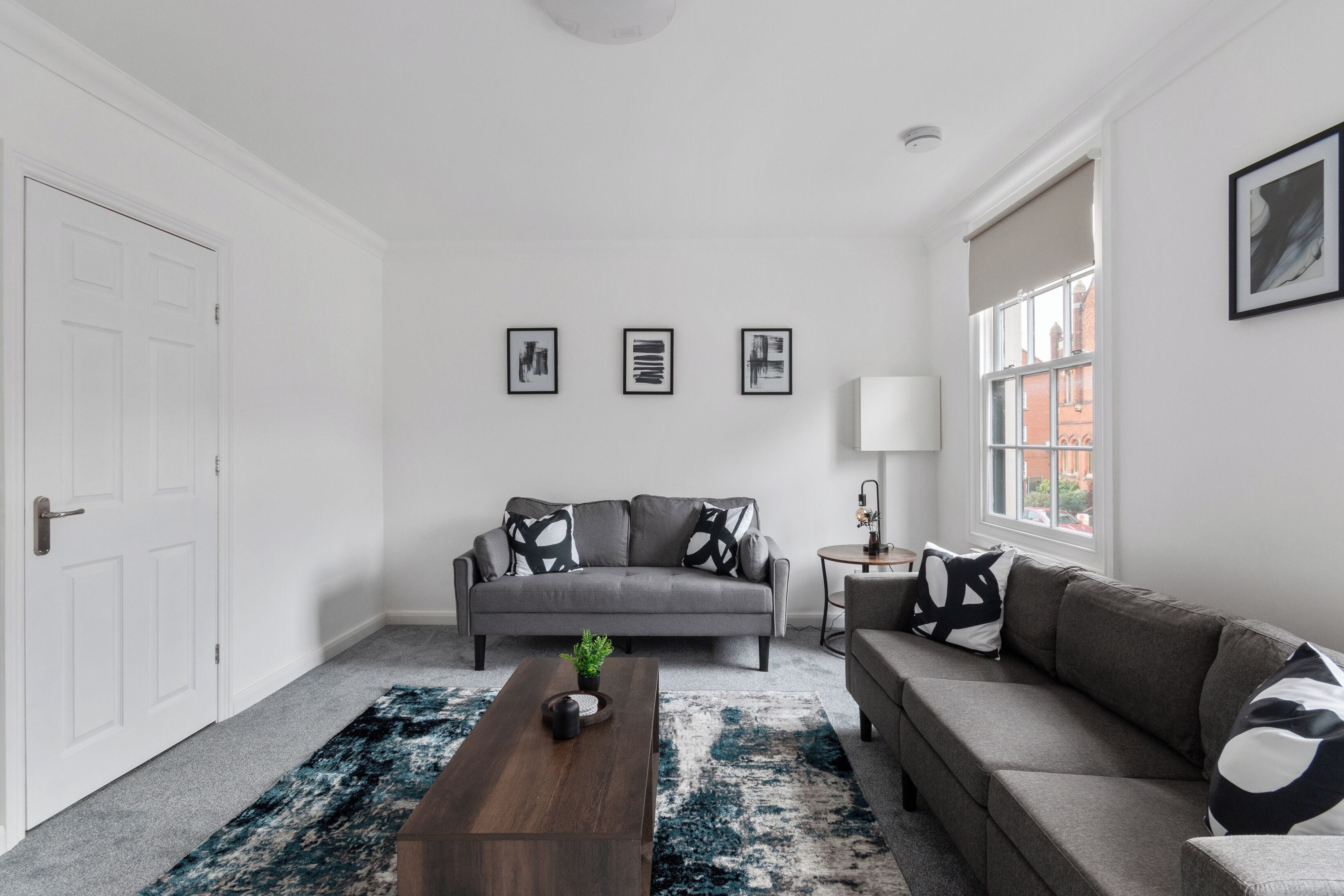 Un salon moderne avec un canapé gris, une table basse en bois et un tapis à motifs. Trois tableaux encadrés ornent le mur, et une fenêtre offre une vue sur les bâtiments.