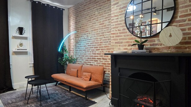 A modern living room with a brick wall, a round mirror, a sofa, a small table, and a fireplace.
