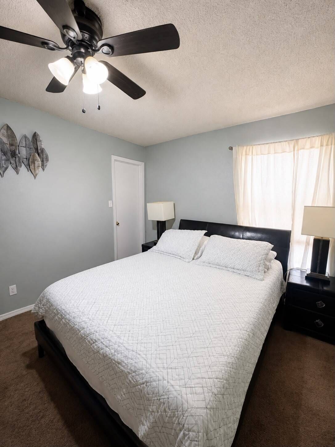 A bedroom with a bed, a ceiling fan, a nightstand, a lamp, and a window with curtains.