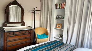A bedroom with a wooden dresser, a mirror, a wicker chair, a bookshelf, and a bed with a striped blanket. - 2BD Garden Hideaway in Lewes (Lewes)