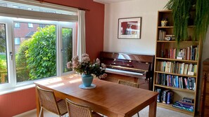 A room with a wooden piano, a bookshelf filled with books, a table with chairs, and a window with a view of greenery. - 2BD Garden Hideaway in Lewes (Lewes)