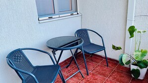A small outdoor seating area with two blue metal chairs and a round table, a window with blinds, and a potted plant. - Nova City Center - 217 (Divjakë)