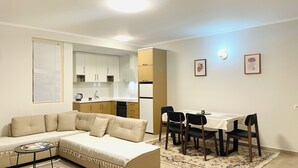A compact living space with a sofa, a dining table with chairs, a kitchen area with cabinets, and a patterned rug. - Nova City Center - 217 (Divjakë)