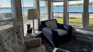 A cozy room with a floral armchair, a dark blue sofa, and a lamp on a small table. Large windows offer a view of a lake and greenery.