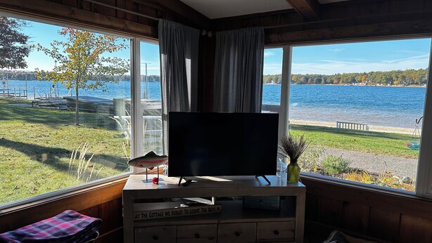 A cozy room with a large window offering a view of a lake, a wooden entertainment unit with a TV, and a pet bed.