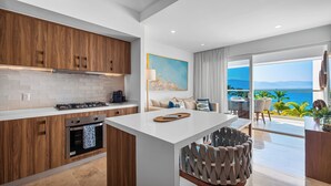 Una cocina moderna con armarios de madera, encimera blanca y una isla central. Hay una zona de comedor con vistas al océano.