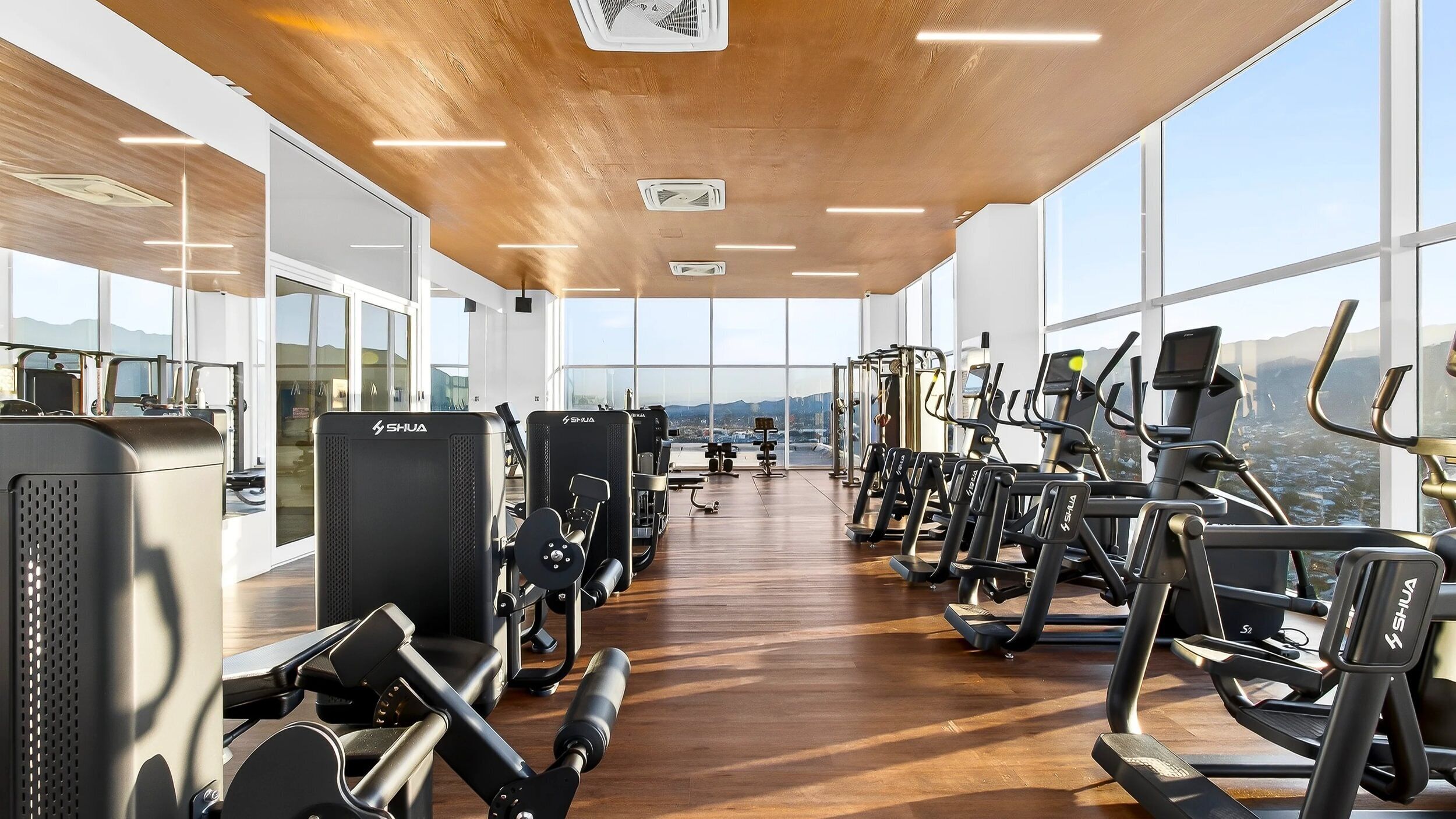 A gym with various exercise equipment, large windows, and a wooden ceiling.