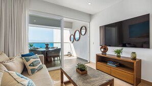 Una sala moderna con sofá, mesa de centro y mueble para televisión. Tiene un balcón con vista al océano.