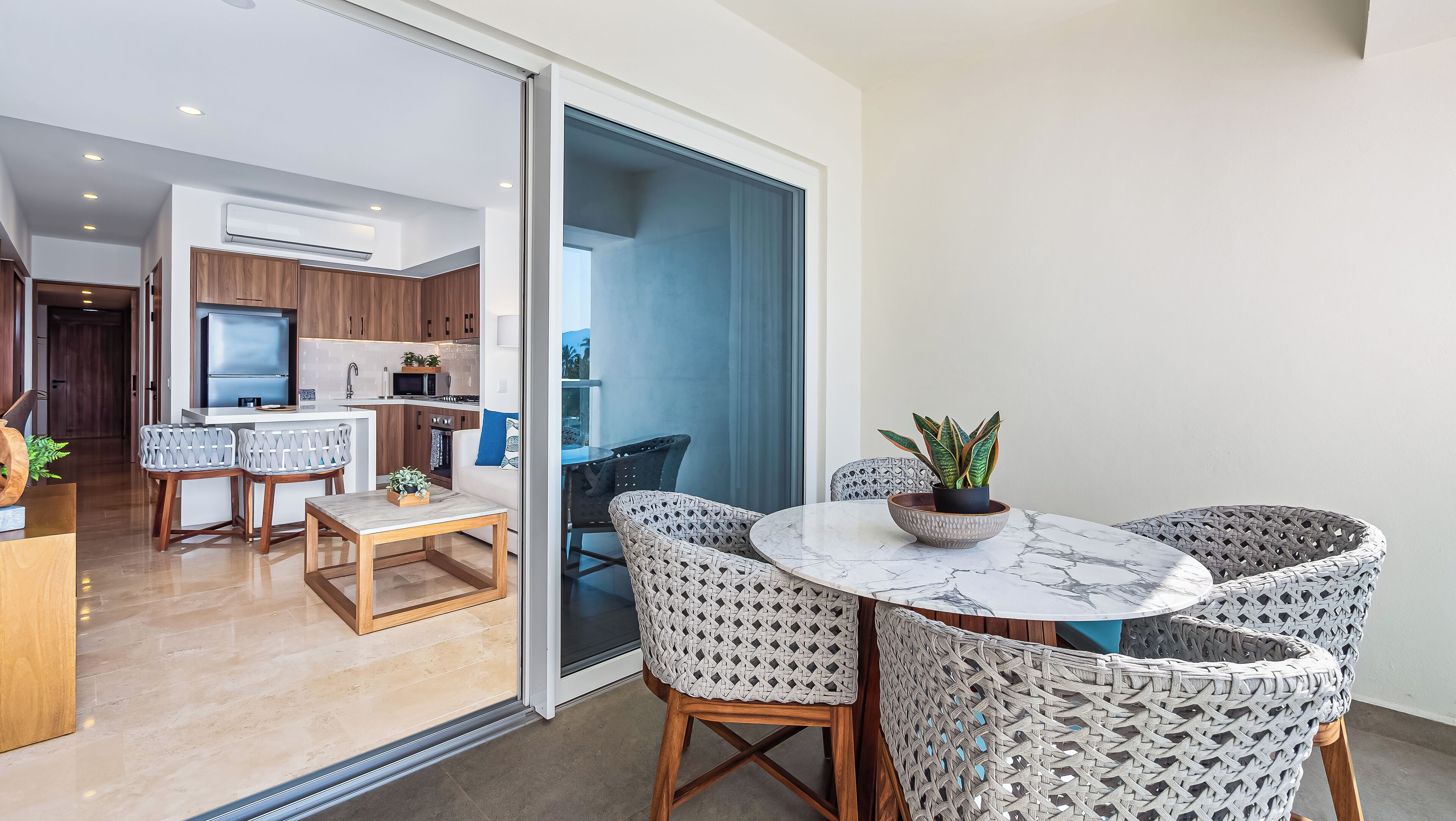 A modern kitchen with a built-in oven and microwave, a dining area with a round table and wicker chairs, and a sliding glass door leading to a balcony.