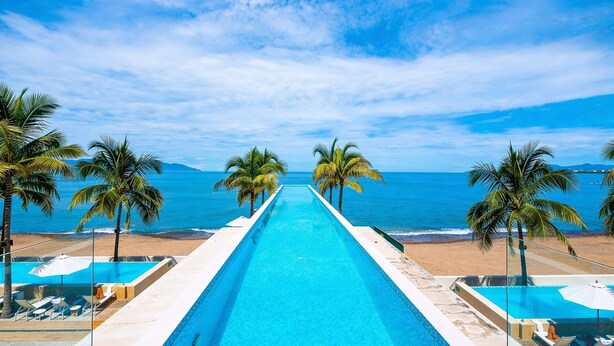 Una piscina larga con agua cristalina azul, rodeada de palmeras y vistas a la playa.