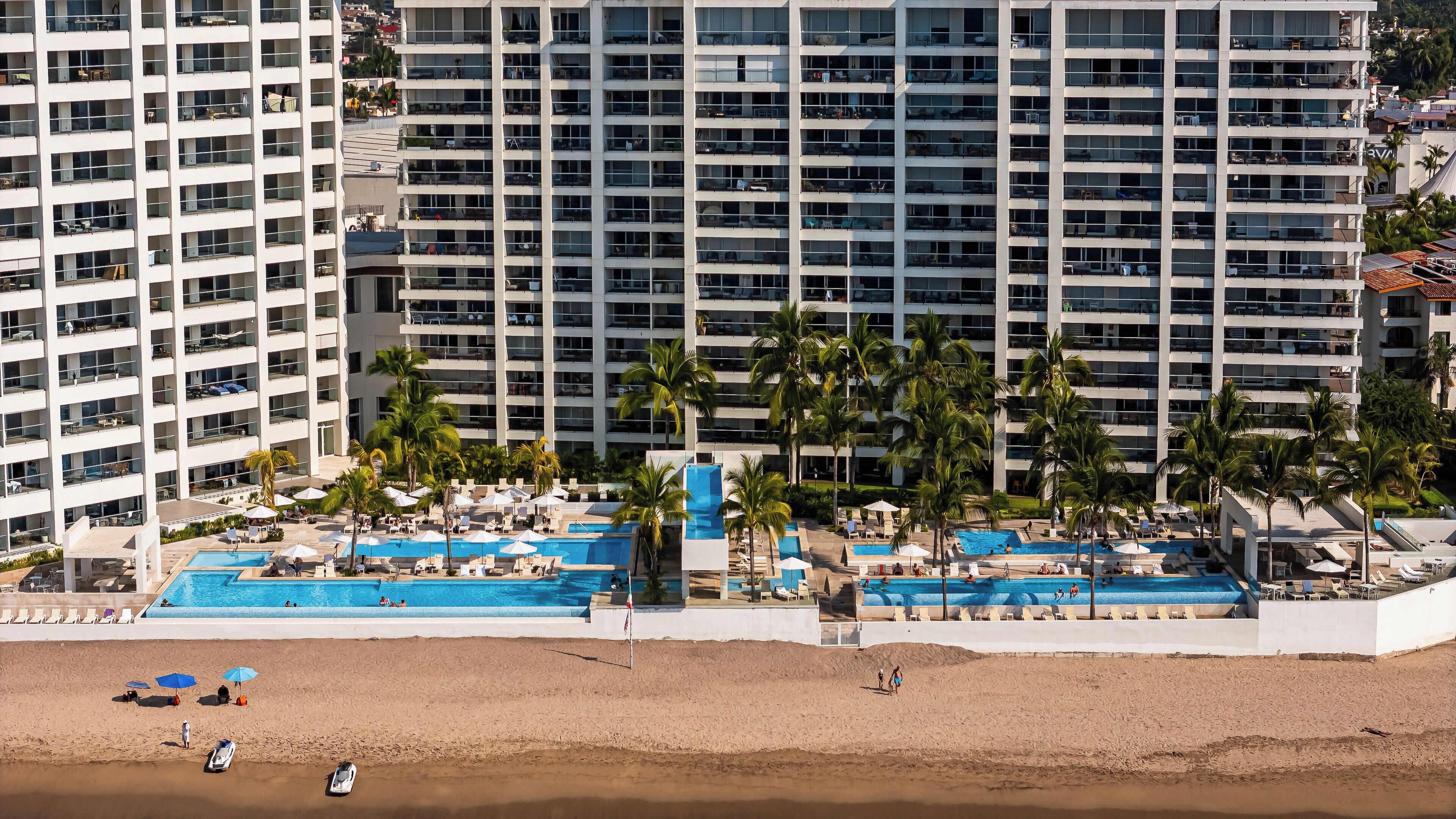 A high-rise hotel with multiple balconies, a pool area with umbrellas, and a beach with sun loungers.