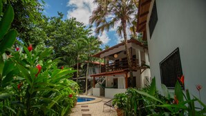 A lush garden with a pool, a wooden pergola, and a white building with a balcony.