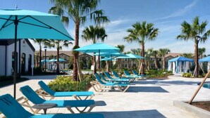 Zwembadterras met blauwe ligbedden en parasols, palmbomen en een heldere lucht.