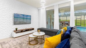 Een moderne woonkamer met een grote televisie, een grijze bank, een salontafel en uitzicht op een zwembad.