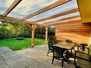 Una pérgola de madera con una mesa y sillas, ubicada en un patio con vista a un jardín.