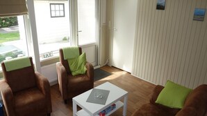A cozy living room with a brown leather sofa, a white coffee table, and two armchairs with green cushions. A window provides a view of the outside. - Bungalow in Sint Maartenszee Near Beach (Sint Maartensvlotbrug)
