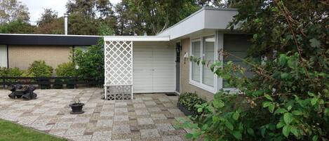 A modern house with a patio area featuring a checkered stone floor, a white lattice fence, and a small garden with potted plants.