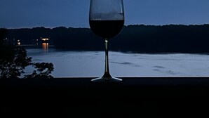 Un verre de vin rouge sur un balcon donnant sur une étendue d’eau, avec une vue lointaine sur la ville et la lune dans le ciel.