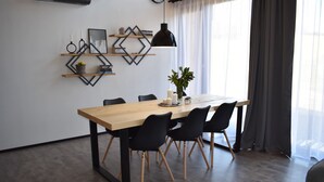 A modern dining area with a wooden table, black chairs, and a pendant light. There are decorative shelves with books and a vase with flowers.