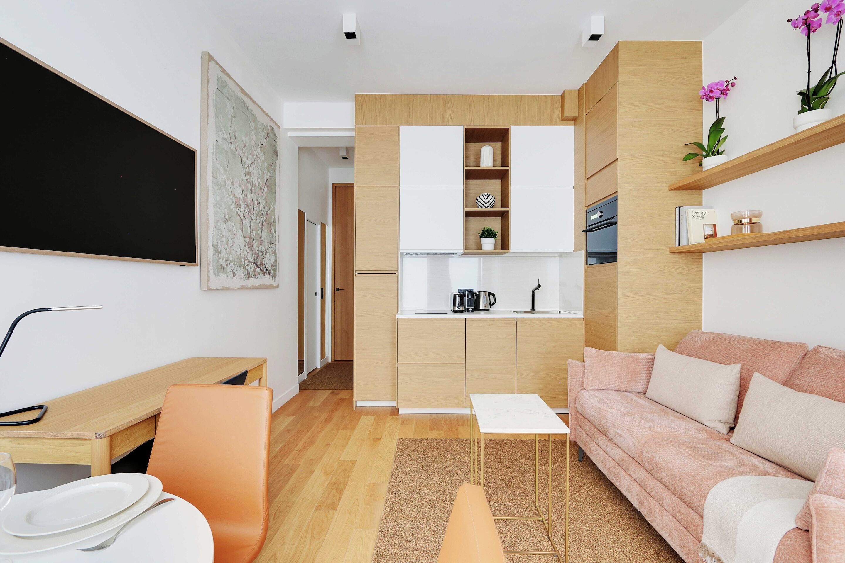 A modern living room with a sofa, a dining table, and a kitchen area with wooden cabinets and a coffee machine.