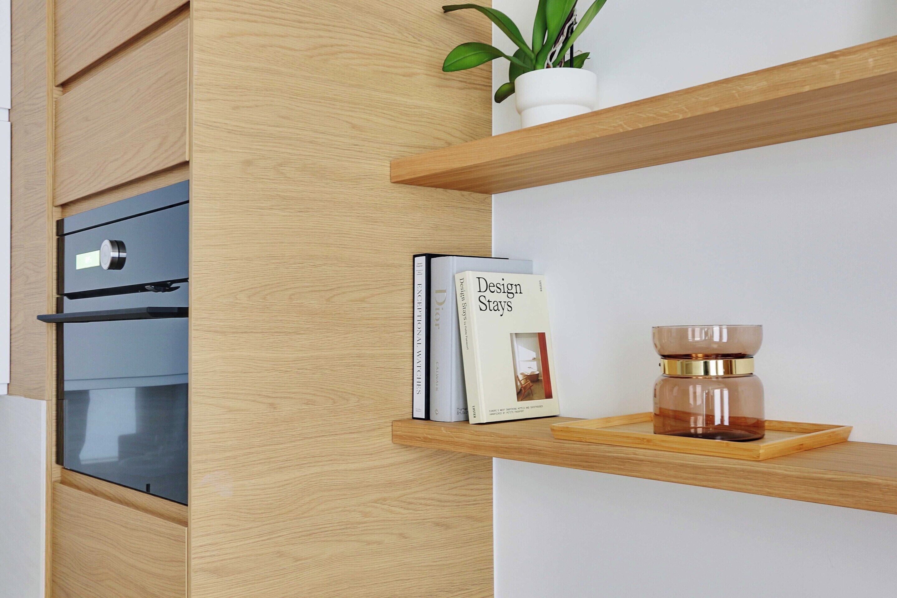 A kitchen with wooden cabinetry, a built-in oven, and a shelf with a book titled 'Design Stays'.