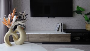 A modern living room with a flat-screen TV mounted on a concrete wall, a wooden cabinet below, and a vase with dried plants on a white table. - Taipei Main Station Warm & Cozy 2B1b Living, Dining, Kitchen (Taipei City)