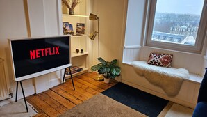 En hyggelig stue med et stort fjernsyn der viser "NETFLIX", en bogreol, en gulvlampe, en potteplante, en vindueskarm med en pude og udsigt til bybilledet.