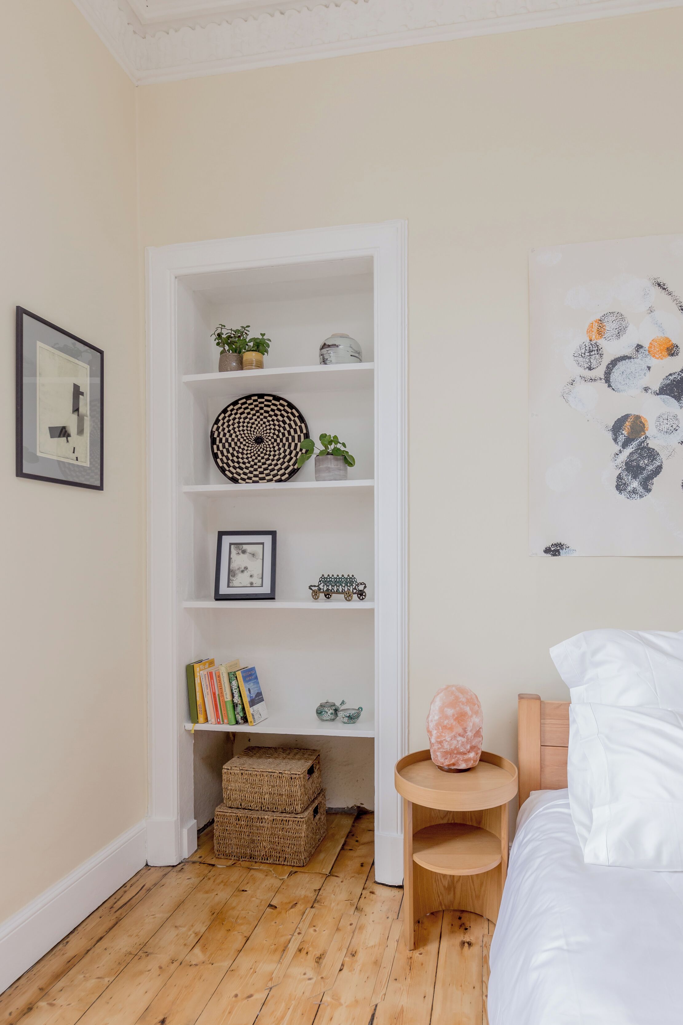 Un dormitorio con piso de madera, una cama blanca, una mesita de noche de madera, una pared blanca con un cuadro enmarcado, una estantería blanca con objetos decorativos, y un cuadro abstracto grande.