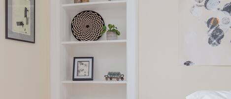 Un dormitorio con piso de madera, una cama blanca, una mesita de noche de madera, una pared blanca con un cuadro enmarcado, una estantería blanca con objetos decorativos, y un cuadro abstracto grande.