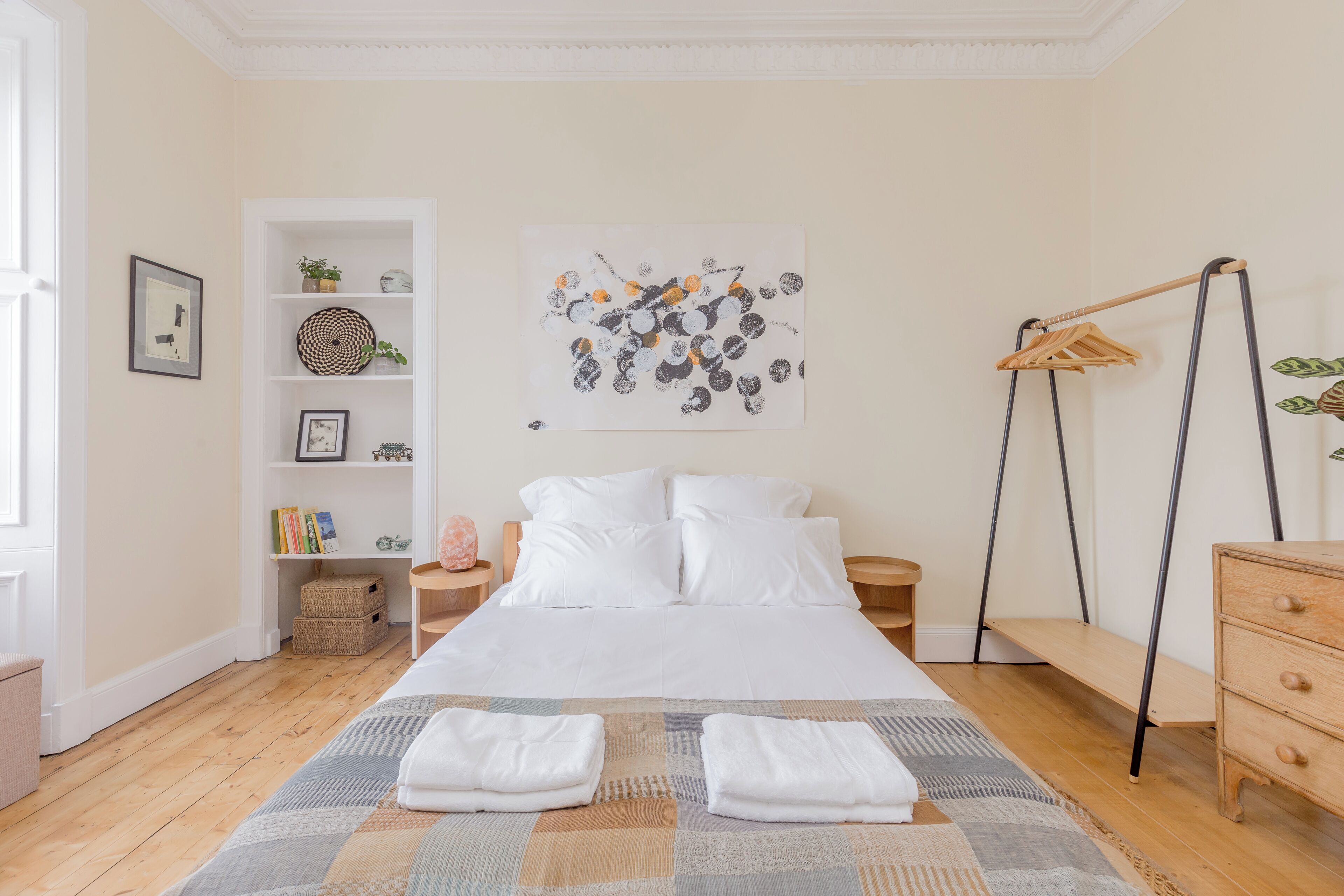 A neatly arranged bedroom with a bed, a hanging clothes rack, a bedside table, and a wall-mounted artwork.