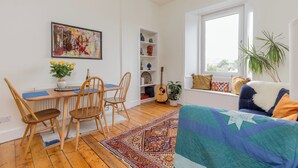 En hyggelig stue med et træbord, stole, et farverigt tæppe, en sofa med puder, en guitar og et vindue med udsigt.