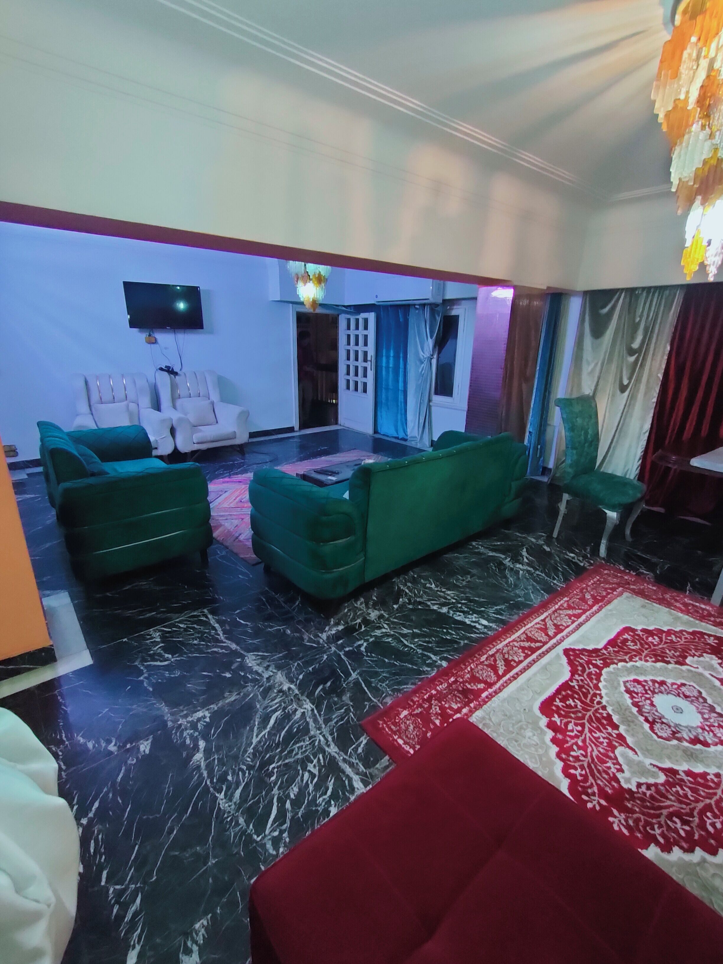 A living room with a large red and white patterned rug, green sofas, and a flat-screen TV mounted on the wall.