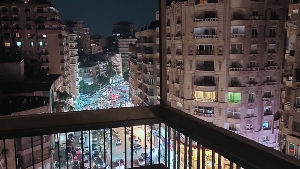A nighttime view from a balcony overlooking a busy street with buildings in the background. - HS STIDUS 1 (Giza)