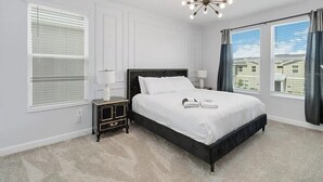 A bedroom with a large bed, a nightstand, a chandelier, and a view of houses through the windows. - Fantastic Home 7BR/5BA with Private Pool • Storey Lake Resort [SL2648] (Kissimmee)