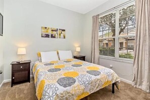 A bedroom with a bed, two bedside tables with lamps, a window with curtains, and a wall with three framed pictures.