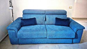 Un sofá azul con dos cojines oscuros en una habitación con una pared blanca lisa y una lámpara de pie.