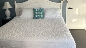 A neatly made bed with a white headboard, a decorative pillow with the text "EAT SLEEP BEACH," and a framed painting above it. - Oceanside St. Rita Hotel Room  (Beach Haven)