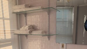 A bathroom with white tiled walls, a toilet, a sink, and shelves with towels. - Oceanside St. Rita Hotel Room  (Beach Haven)