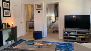Una sala moderna con un televisor de pantalla plana, una mesa de centro con libros, un otoman azul y una estantería.