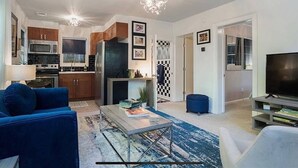 Una sala de estar moderna con un sofá azul, una mesa de centro y un televisor. Cuenta con una zona de cocina con armarios y electrodomésticos.