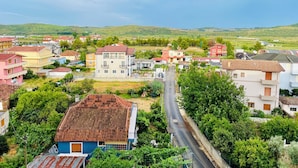A residential area with a variety of houses, a main road, and green hills in the background. - Adora City Center Divjake (Divjakë)