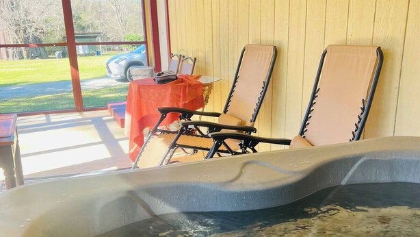A hot tub with two lounge chairs, a table with a red tablecloth, and a view of a grassy yard and parked car.