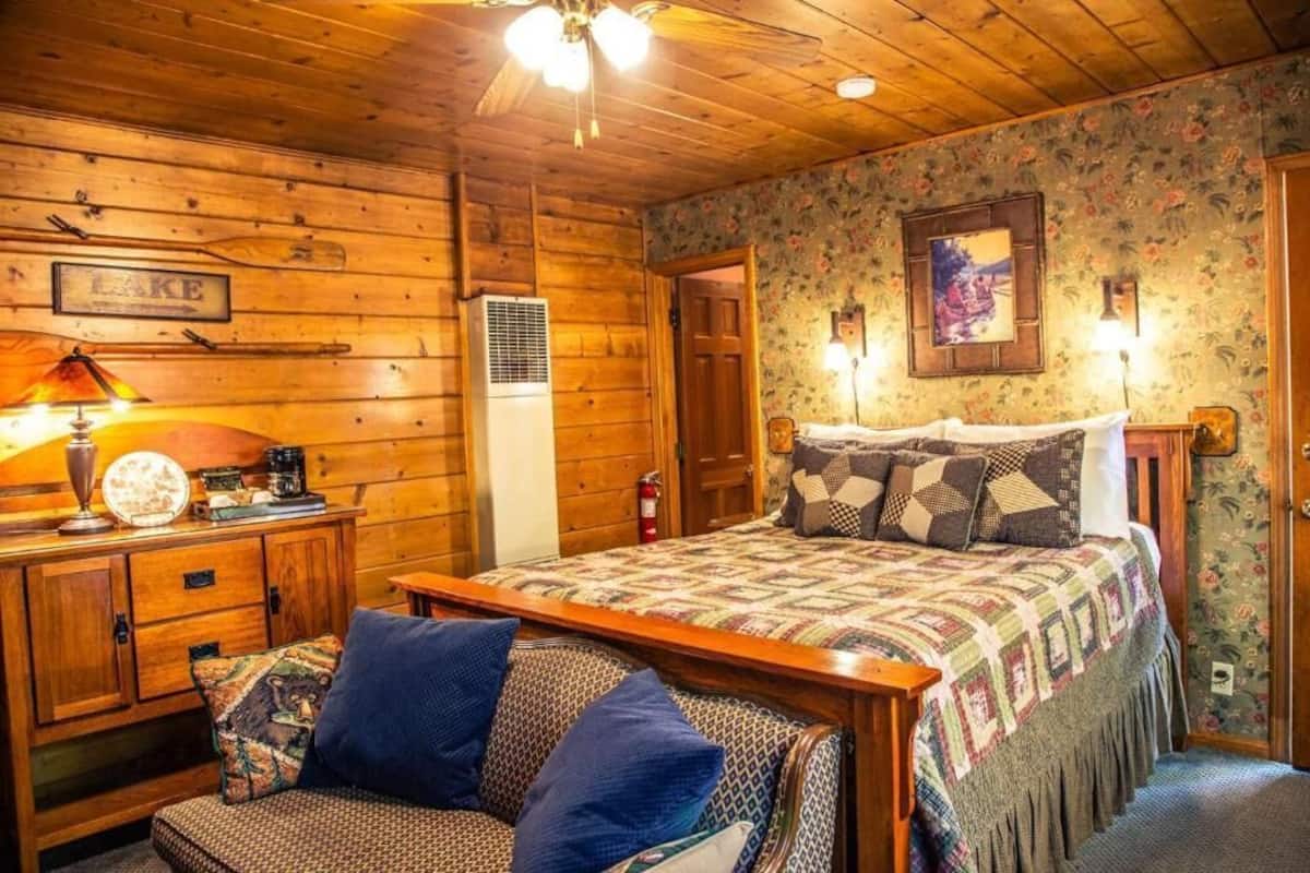 Habitació d'estil rústic acollidora amb sostre de fusta, un llit amb col·cha estampada, un sofà amb coixins blaus, un taulell de nit de fusta i una làmpada penjada a la paret.