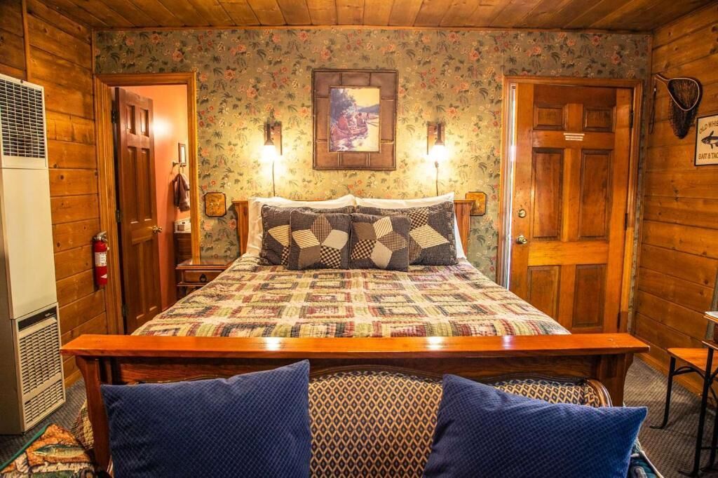 A wooden cabin room with a bed, two wall-mounted lights, a fire extinguisher, and a framed picture on the wall.