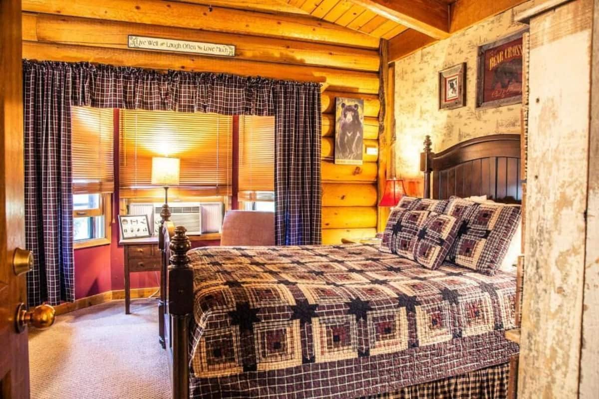 Un dormitori acollidor d'estil cabana de fusta, amb un llit gran, cortines de viqueta i un amb motlle de fusta.