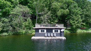 A houseboat with a deck and railing, surrounded by water and trees. - Lake of Bays Getaway -Waterfront Cottage with Scenic Rooftop Boathouse (Dwight)