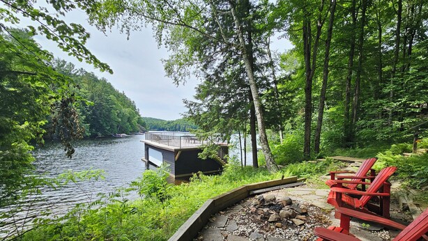 A lakeside deck with red Adirondack chairs, a dock extending into the water, and a forested backdrop. - Lake of Bays Getaway -Waterfront Cottage with Scenic Rooftop Boathouse (Dwight)