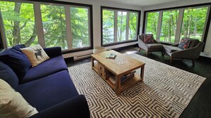 A modern living room with a blue sofa, a wooden coffee table, and a patterned rug. Large windows provide a view of trees outside. - Lake of Bays Getaway -Waterfront Cottage with Scenic Rooftop Boathouse (Dwight)